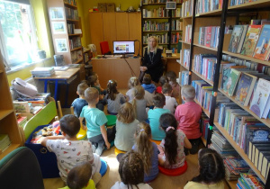 Dzieci siedzą pomiędzy regałami książek, słuchają wiersza czytanego przez Panią Małgorzatę Hofmann.